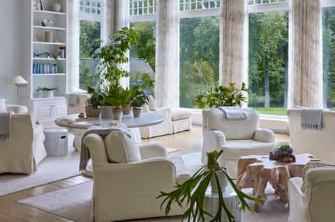 Bright sunroom with white upholstered furniture, potted plants, large windows, built-in shelving, and nature views
