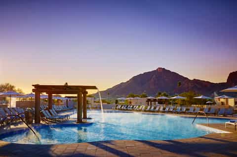 Resort pool at dusk with mountain backdrop, pergola structures, lounge chairs, and blue umbrellas