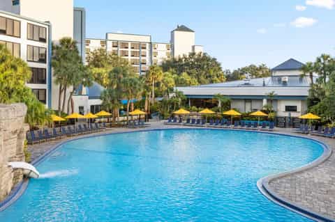 Curved resort pool with yellow umbrellas, lounge chairs, and palm trees surrounded by multi-story hotel buildings