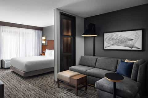 Modern hotel suite with king bed, gray sofa, dark accent wall, and contemporary furniture and lighting