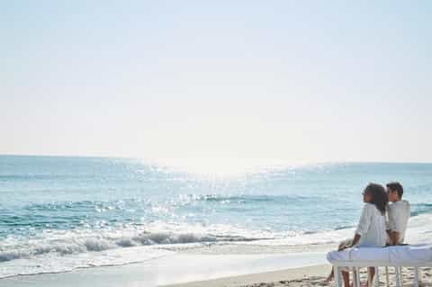 Couple sitting on white bench on sandy beach watching turquoise ocean waves at sunset