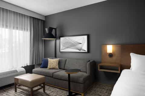 Modern hotel suite with gray sofa, queen bed, wooden nightstand, and contemporary artwork
