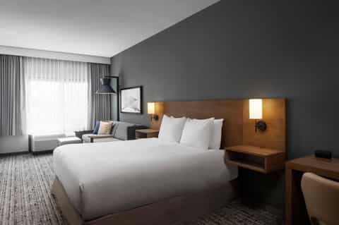 Modern hotel bedroom with queen bed, wooden headboard with integrated lighting, gray walls, and sitting area