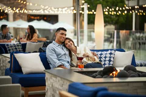 Couple enjoying drinks by outdoor fireplace with string lights and comfortable seating