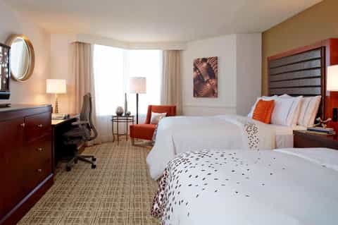 Hotel bedroom with two queen beds, dark wood headboards, orange accent chair, and work desk