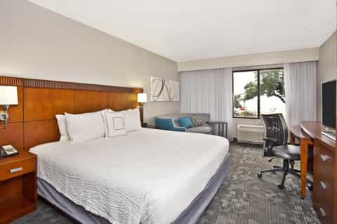 Contemporary hotel room with queen bed, wood headboard, sofa, work desk, and two large windows