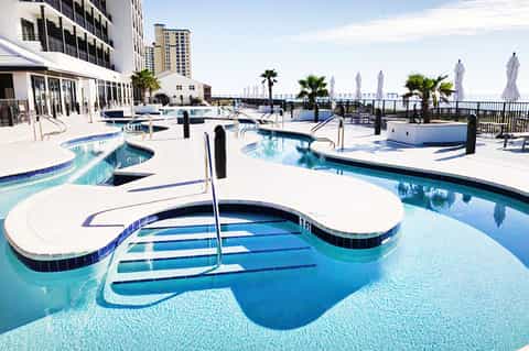Rooftop pool deck with multiple pools, umbrellas, ocean view, and high-rise hotel building