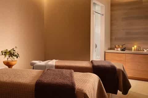 Spa treatment room with massage tables, warm lighting, wooden dresser, and plants
