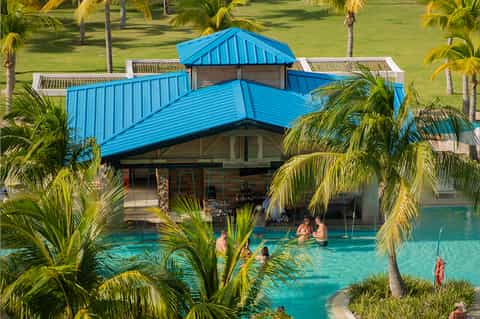 Tropical resort pool with blue roof pavilion, palm trees, and guests swimming in turquoise water