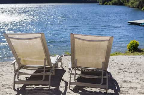 Two lounge chairs facing a sparkling lake with a small table between them on sandy beach