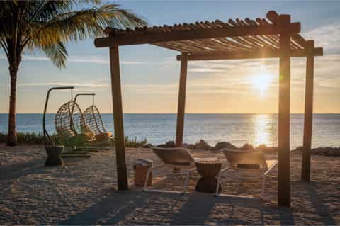 Beach sunset view with wooden pergola, hanging egg chair, lounge chairs, and palm tree at dusk
