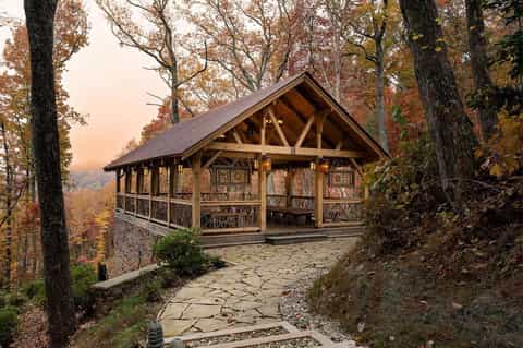 Rustic wooden pavilion with stone patio nestled among autumn trees on hillside at dusk