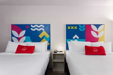 Modern hotel room with two queen beds, colorful geometric headboard artwork, and red accent pillows