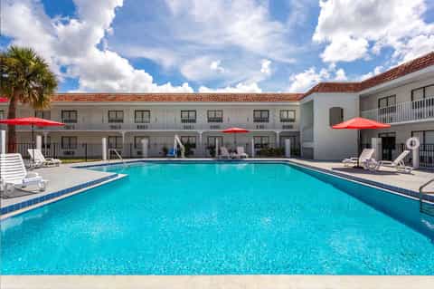 Outdoor pool surrounded by white hotel buildings, lounge chairs, and red umbrellas under clear sky