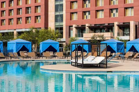 Resort pool with blue cabanas, tan loungers, modern red brick building, and white cabana bed in foreground