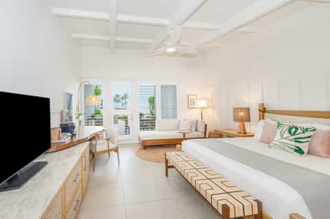 Bright hotel suite with queen bed, sitting area, TV, and ocean-view balcony doors