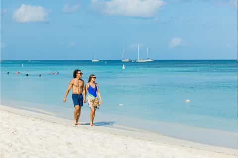 Couple walking on white sand beach with sailboats on clear turquoise water