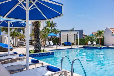 Resort swimming pool with blue umbrellas, loungers, white pergola structures, and palm trees under clear sky