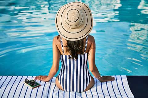 Woman in striped swimsuit and wide-brimmed hat sitting poolside with phone