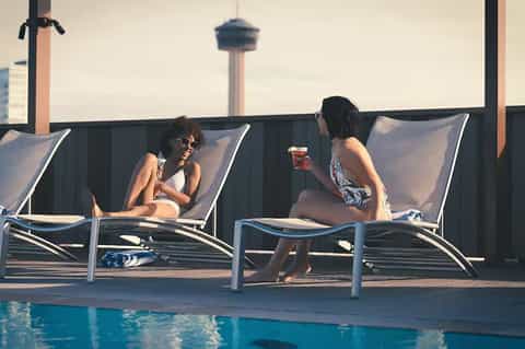 Two women relaxing in lounge chairs by a turquoise pool with city skyline in background