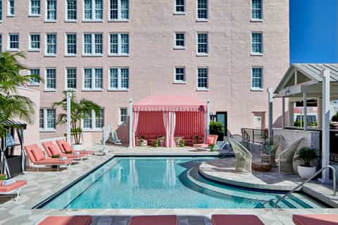 Hotel courtyard with rectangular pool, lounge chairs, pink cabanas, and modern building