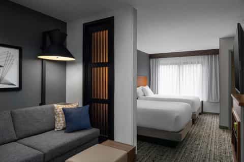 Contemporary hotel room with gray sofa, black partition, and view into bedroom with white linens