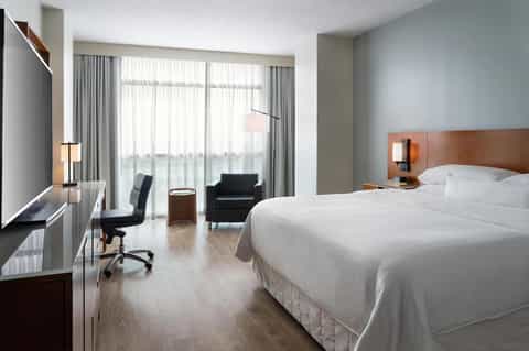 Modern hotel bedroom with king bed, work desk, armchair, and floor-to-ceiling windows
