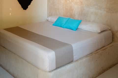 Minimalist hotel room with white bed, turquoise pillow, and concrete walls