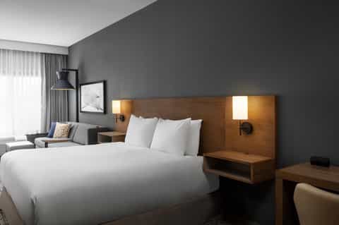 Contemporary hotel bedroom with wood headboard, white bedding, gray walls, and sitting area