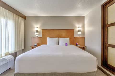 Modern hotel bedroom with wood headboard, white bedding, elegant wall sconces, and bright window light