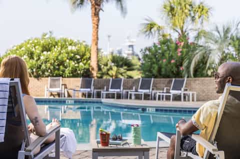 Couple relaxing poolside on lounge chairs with drinks and palm trees in background