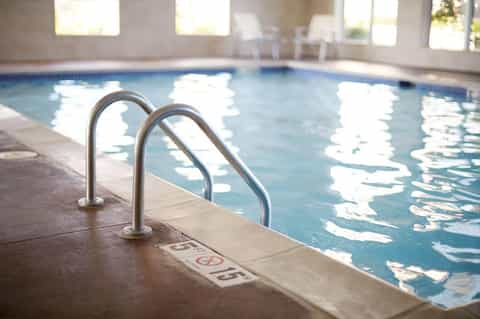 Indoor hotel pool with metal ladder railing, bright natural light streaming through windows, white chairs visible