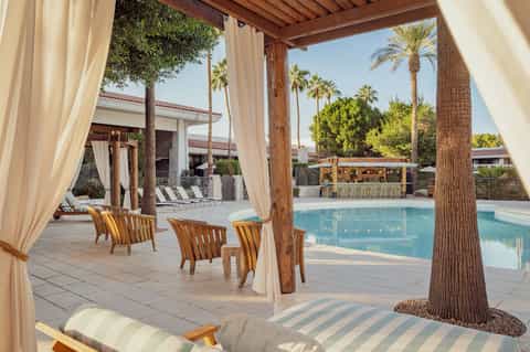 Poolside pergola with cream curtains, wooden lounge chairs, and tropical palm surroundings