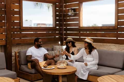 Resort patio with three guests enjoying drinks on gray sofa under wooden pergola with ocean view