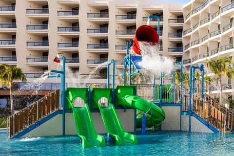 Colorful water park with green slides, red bucket dumper, and blue poles in turquoise pool with tall hotel buildings