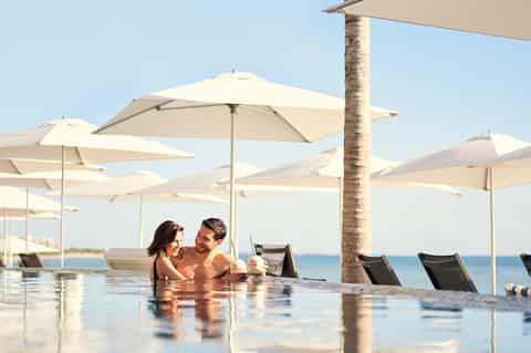 Couple relaxing in infinity pool with ocean views, palm trees, and white umbrellas in background