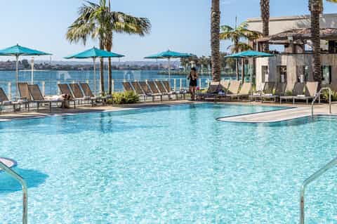 Resort pool with turquoise water, lounge chairs, teal umbrellas, palm trees, and waterfront view