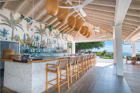 Tropical beach bar with white columns, woven pendant lights, wooden stools, and ocean views