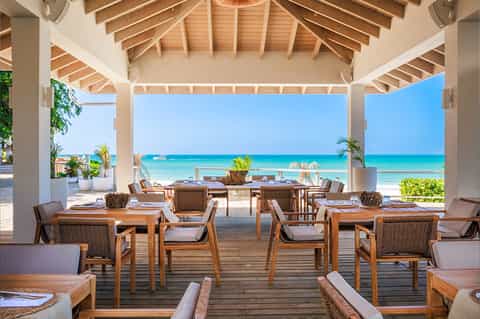Beachfront dining pavilion with wooden tables, rattan chairs, and clear ocean view under white roof