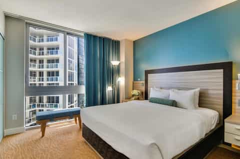 Modern hotel bedroom with teal accent wall, king bed, floor-to-ceiling windows overlooking city skyline