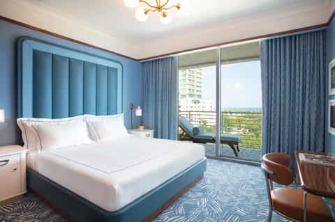 Luxury hotel bedroom with blue upholstered headboard, white linens, balcony view, and modern furnishings