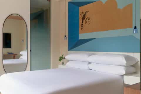 Modern hotel bedroom with white bedding, arched mirror, blue and tan wall mural, and pendant lighting