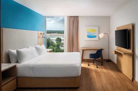 Hotel bedroom with blue accent wall, queen bed, wooden desk, TV, and panoramic window overlooking river landscape