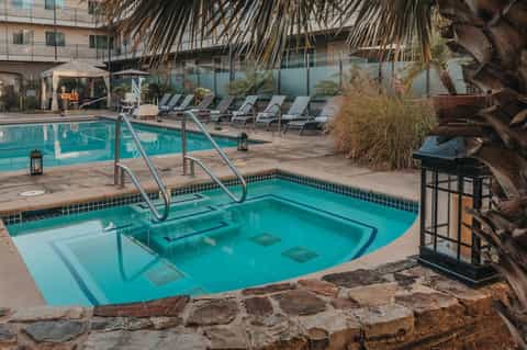 Resort pool deck with turquoise swimming pools, spa area, lounge chairs, palm trees, and multi-story building in background