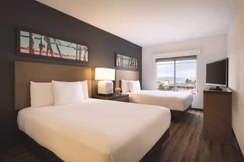 Hotel room with queen bed, wood headboard, palm tree artwork, television, and coastal window view