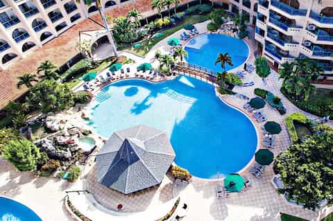 Aerial view of resort with curved blue pools, palm trees, and surrounding apartment buildings
