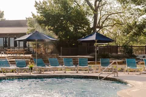 Outdoor resort pool with lounge chairs, blue umbrellas, wooden deck, trees, and gazebo areas