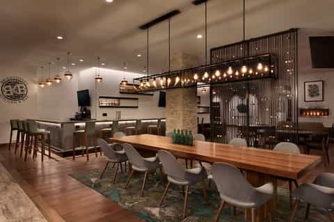 Modern hotel restaurant with wooden dining table, pendant lighting, and decorative bar area