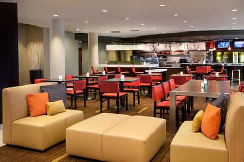Modern hotel common area with cream seating, orange accents, dining tables, and open kitchen concept