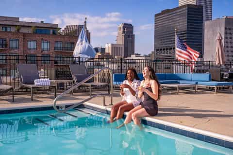 Two women sitting poolside at rooftop pool with city skyline and American flags visible